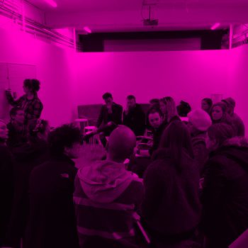 A Group of people sitting in a room with one person writing on a whiteboard, the image is over-layed with a pink filter