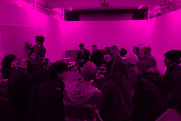 A Group of people sitting in a room with one person writing on a whiteboard, the image is over-layed with a pink filter